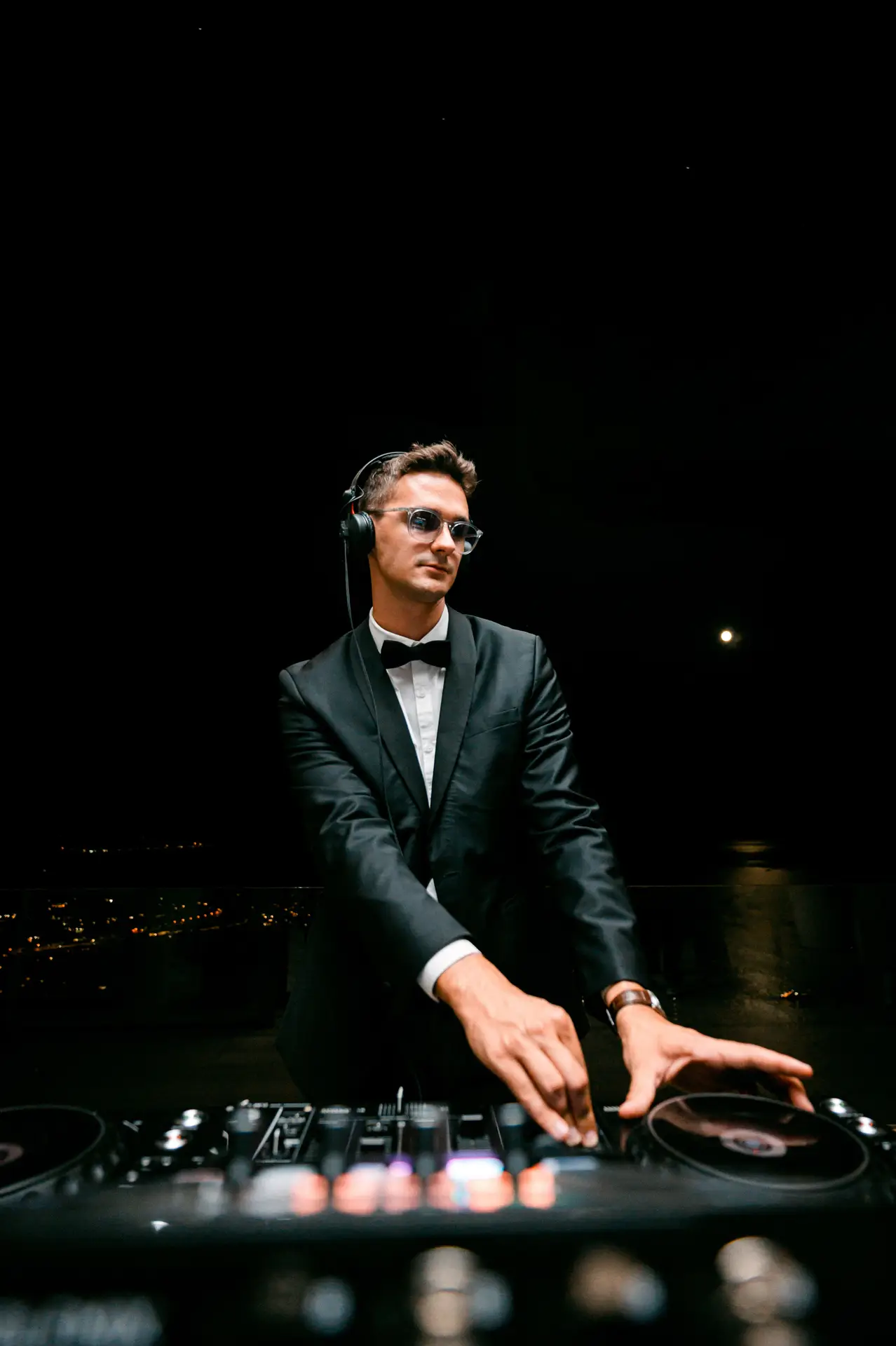 DJ in a black tuxedo and bow tie mixing music on a deck at a night event on the French Riviera
